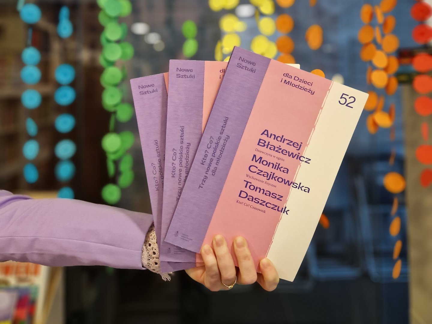 Trzy numery Nowych Sztuk trzymane w dłoni na tle kolorowych girland.