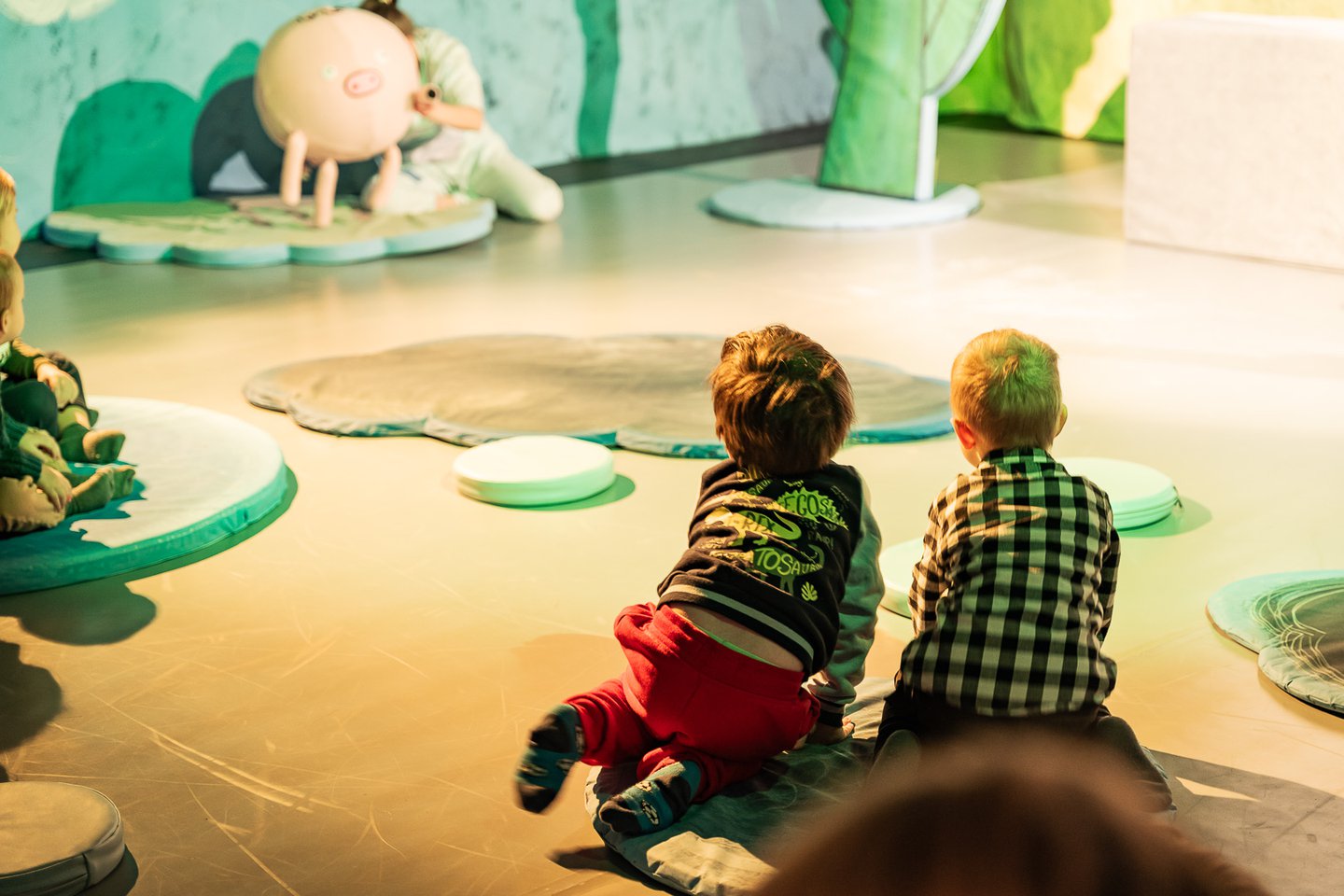 Dwoje małych chłopców siedzi na podłodze i spogląda na znajdującą się na drugim końcu pomieszczenia świnkę - lalkę teatralną.
