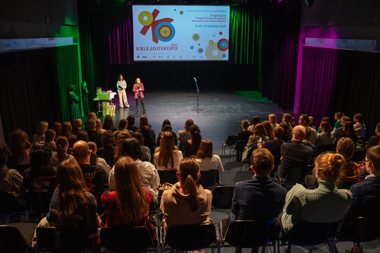Ciemna sala, wypełniona publicznością. Na scenie dwie kobiety i mikrofon. W tle duży ekran z kolorową grafiką i napisem: Oko Kalejdoskopu.