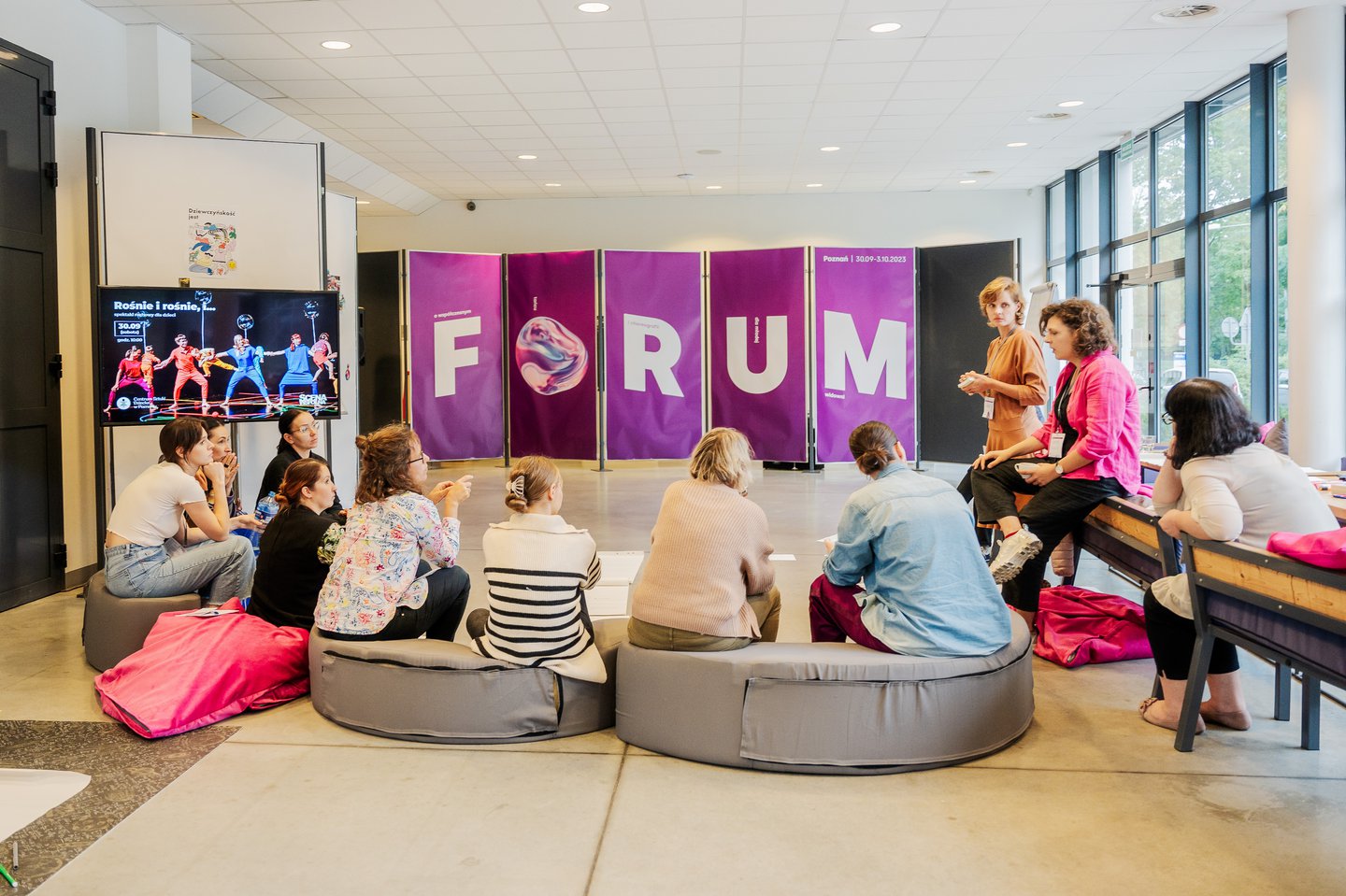 Kilka osób siedzi na miękkich ławach, obok nich stoją kolejne osoby, wszyscy rozmawiają ze sobą. Przed nimi, na stelażach, wiszą duże, fioletowe plakaty z literami tworzącymi napis: Forum.