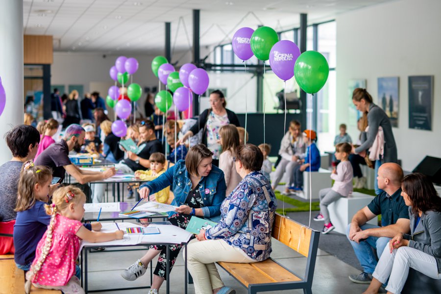 Oszklone foyer Sceny Wspólnej pełne ludzi. Dorośli oraz dzieci siedzą na ławkach, poduszkach, rozmawiają ze sobą. Pomiędzy nimi kolorowe balony z napisem: Scena Wspólna.