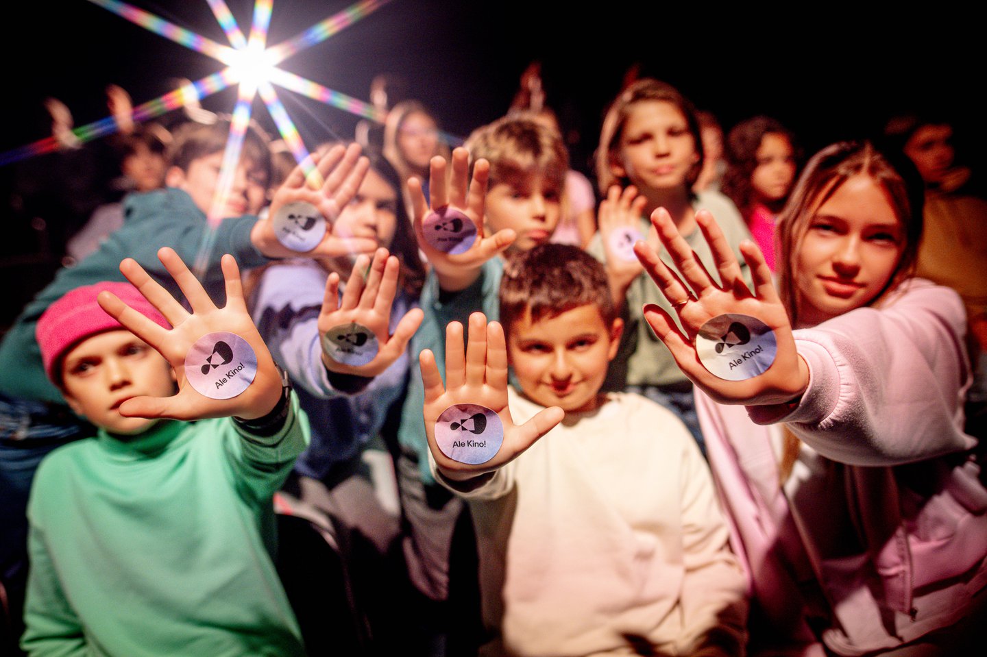 Grupa nastolatków siedzi w grupie na krzesłach na widowni. Wyciągają przed siebie otwarte dłonie, na których mają naklejone naklejki z napisem: Ale Kino!