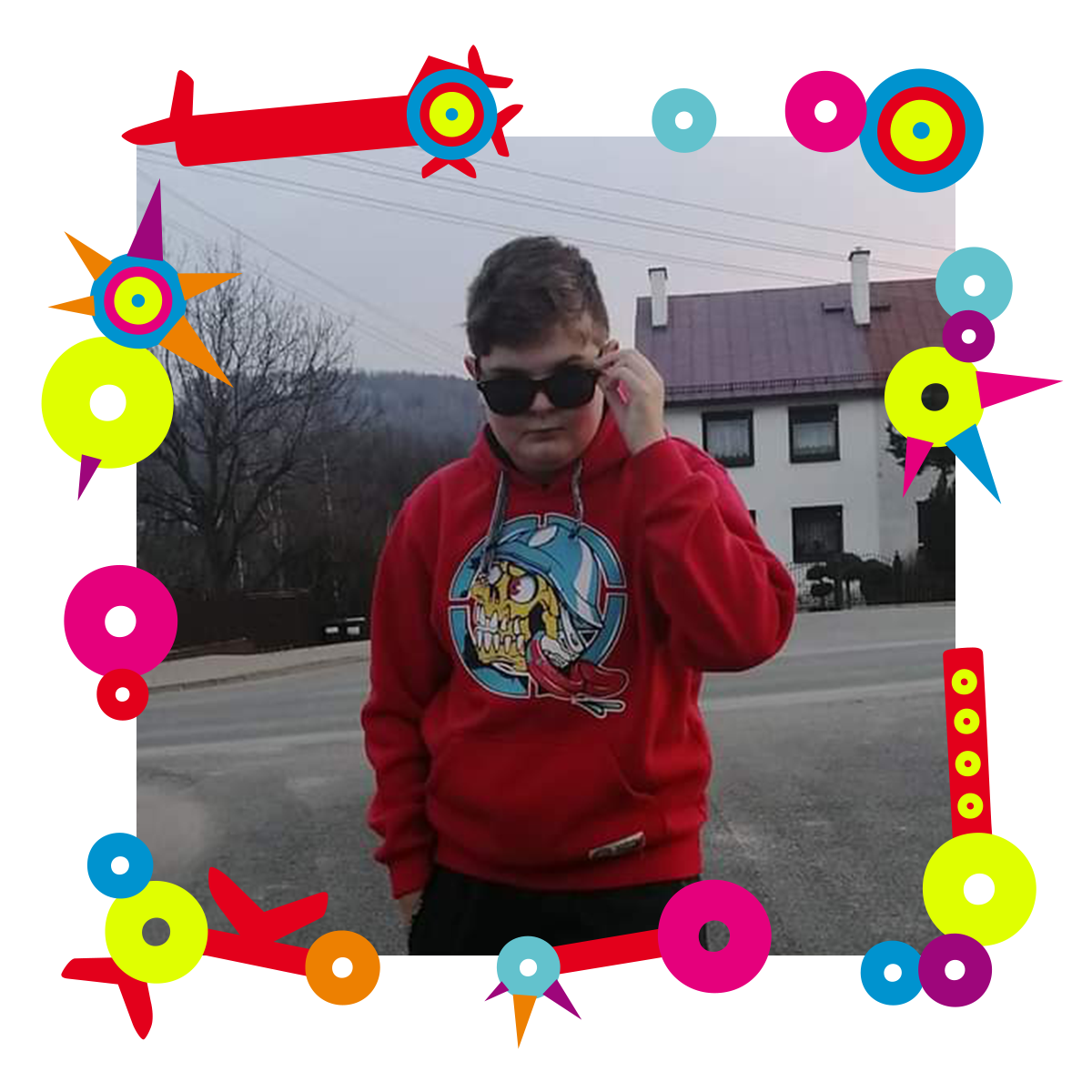 Na zdjęciu chłopiec stojący przed domem. Ma na oczach okulary przeciwsłonecznie, które trzyma lewą ręką. Ubrany jest w bluzę z kapturem i dresowe spodnie. Dookoła zdjęcia kolorowa ramka.