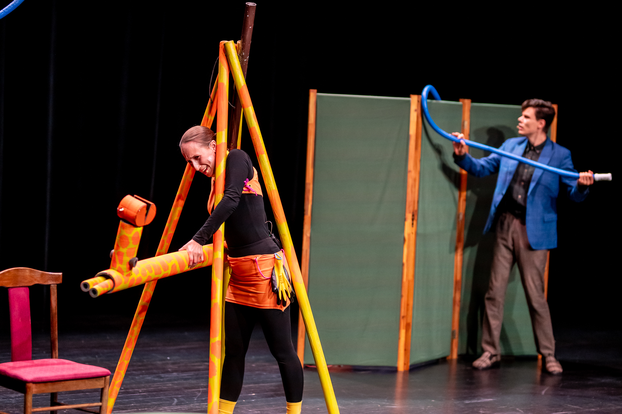 Na scenie aktor i aktorka. Aktor stoi przy rozłożonym parawanie, trzyma w rękach niebieską, długą rurkę. Aktorka stoi pod żółto-pomarańczowymi tubami przypominającymi długie nogi żyrafy i trzyma w rękach kolejną tubę wyglądającą jak szyja z głową żyrafy.