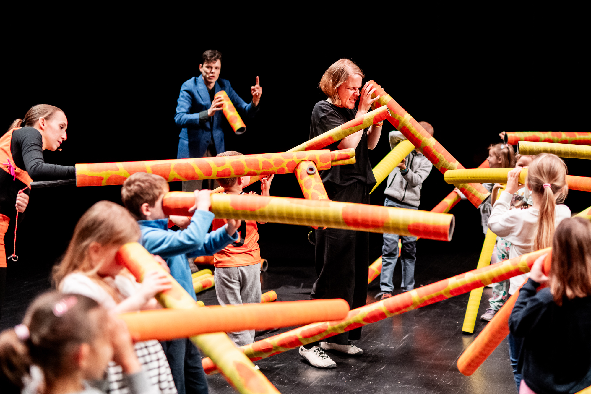 Grupa dzieci i kilkoro dorosłych patrzy przez długie, żółte w pomarańczowe plamy, tuby.