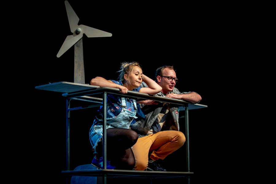 Kobieta i mężczyzna kucają na podwyższeniu, w elementach scenografii, w tle widać drewniany wiatrak.
