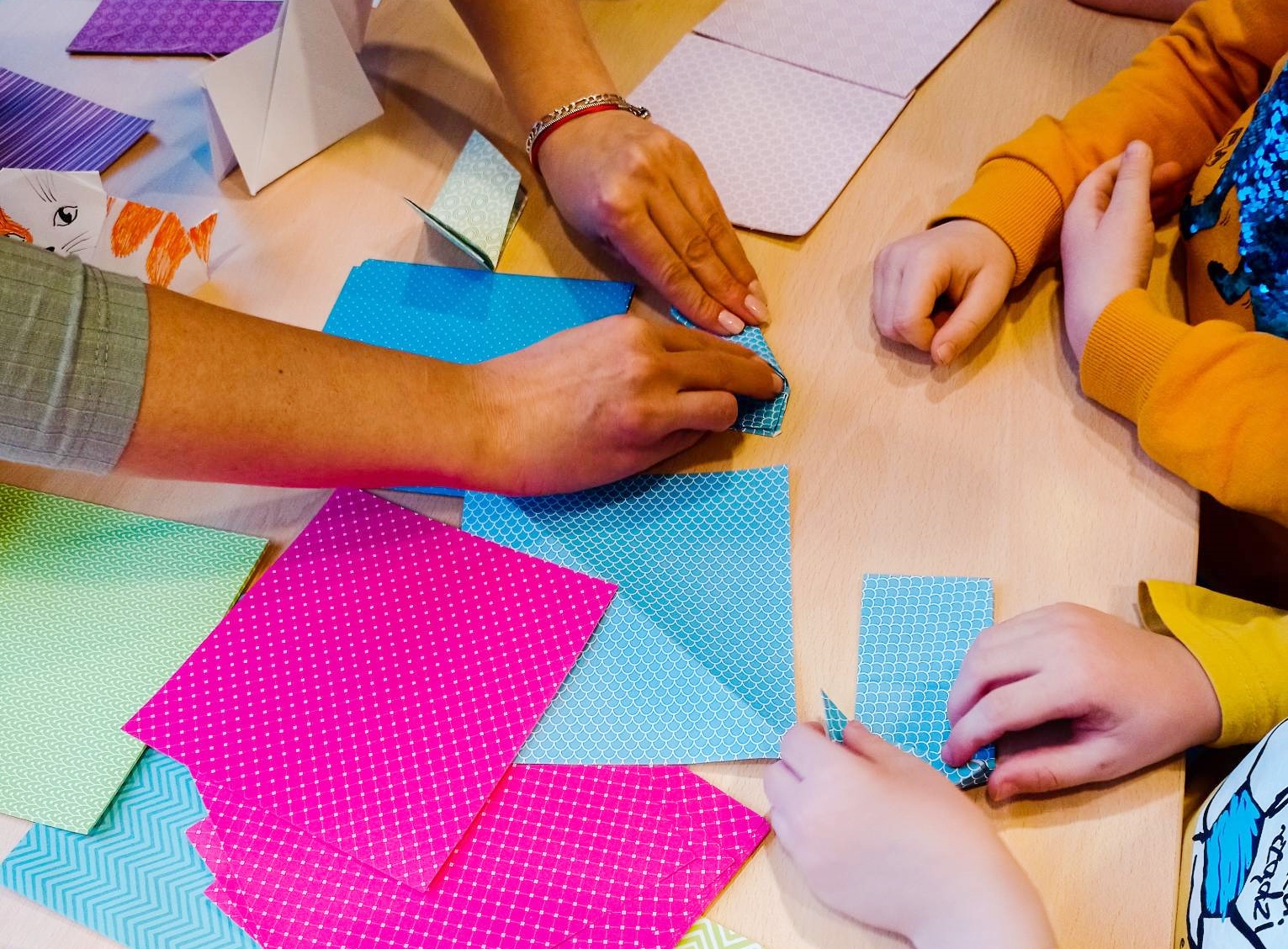 Na zdjęciu trzy pary dłoni składających kształty z kolorowych kartek do origami.