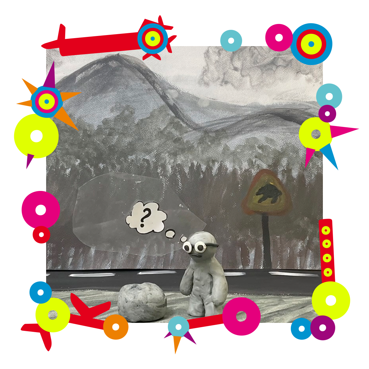Na zdjęciu postać z plasteliny. Stoi obok narysowanego znaku drogowego. Za nim namalowane góry. Dookoła zdjęcia kolorowa ramka.