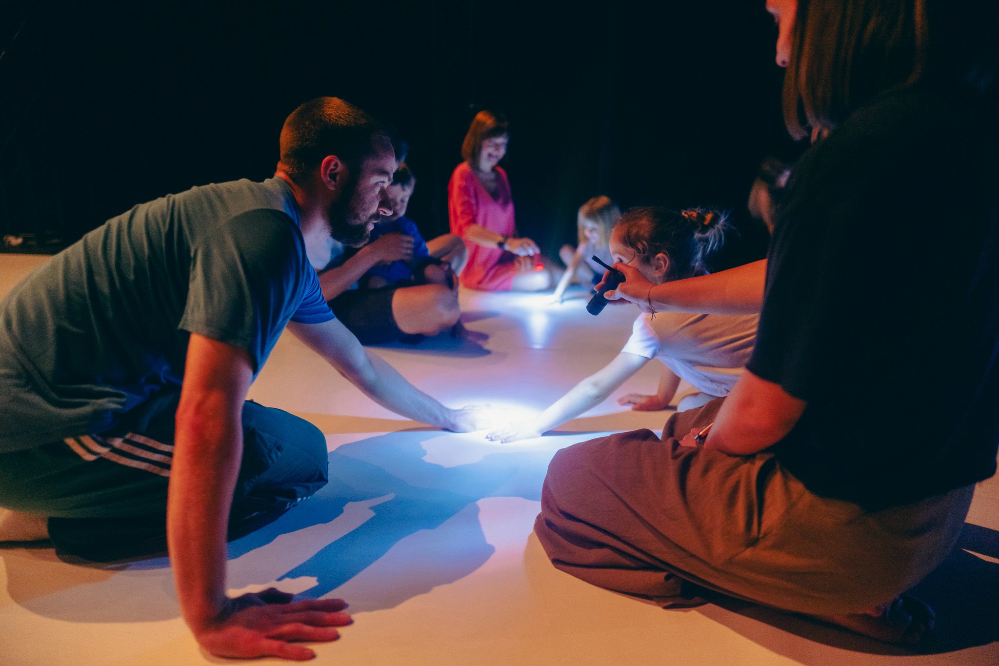 Dzieci i osoby dorosłe siedzą na podłodze. Jest dość ciemno, na środku mały punkt światła z latarki.