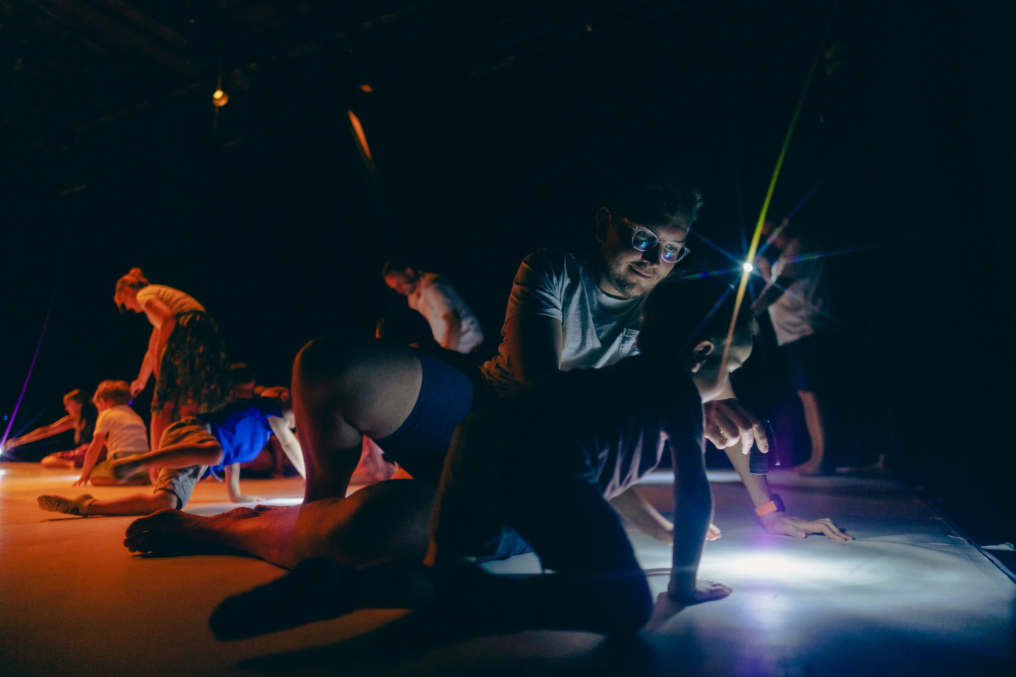 W dość ciemnej sali dzieci i dorośli siedzą na podłodze. Między nimi mały punkt światła z latarki.