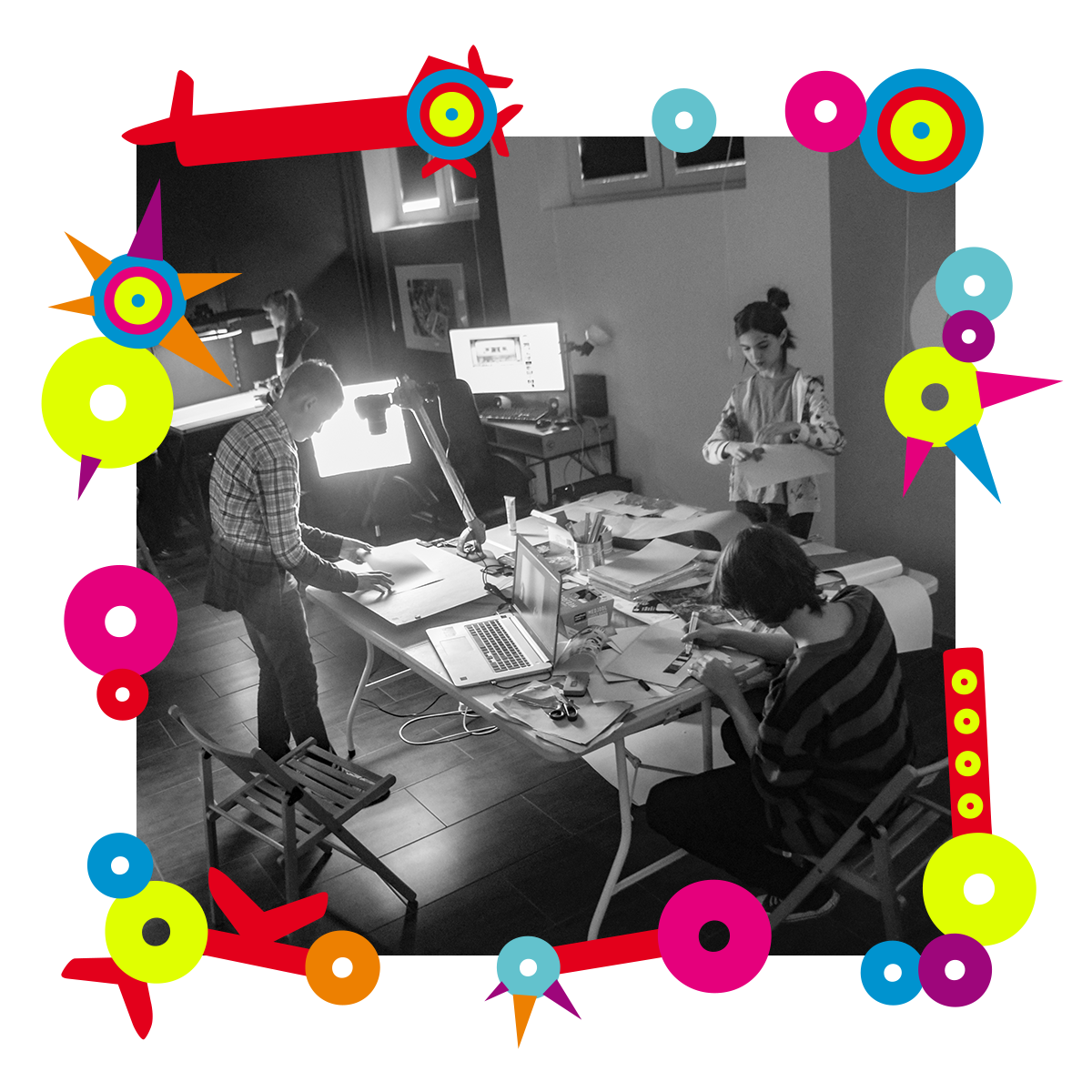 Na zdjęciu pracownia artystyczna. Trzy osoby nachylają się nad stołem, nad którym leżą kartki, komputery. Jedna z nich rysuje coś. Dookoła zdjęcia kolorowa ramka.