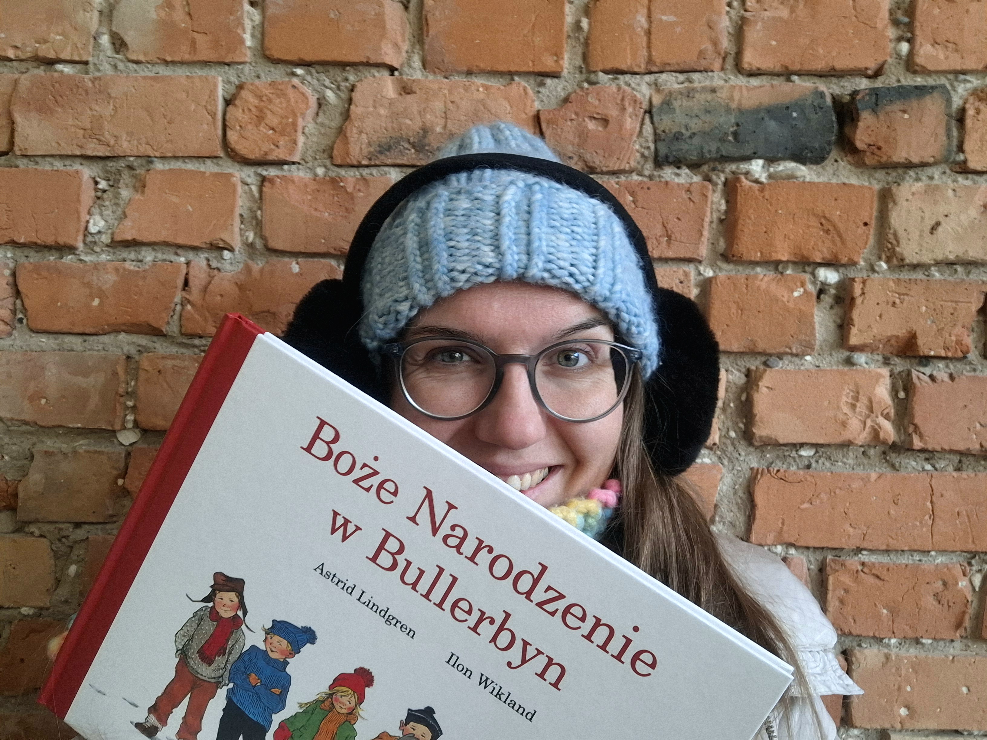 Uśmiechnięta kobieta w czapce i nausznikach trzyma przed sobą książkę: Boże Narodzenie w Bullerbyn.