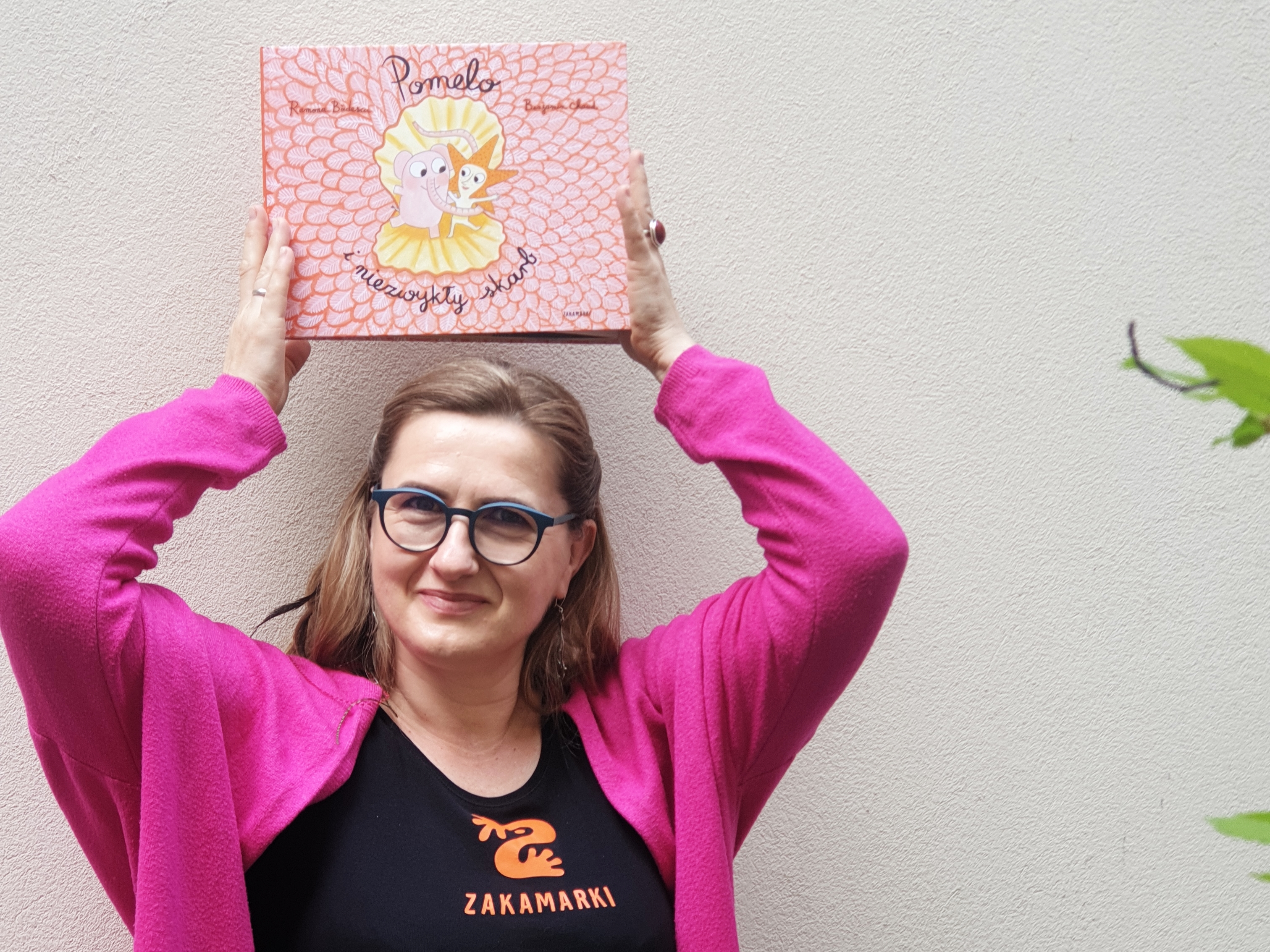 Uśmiechnięta kobieta unosi nad głową książkę „Pomelo i niezwykły skarb". Kobieta ma na sobie różowy sweter i czarną koszulkę z logo Wydawnictwa Zakamarki.