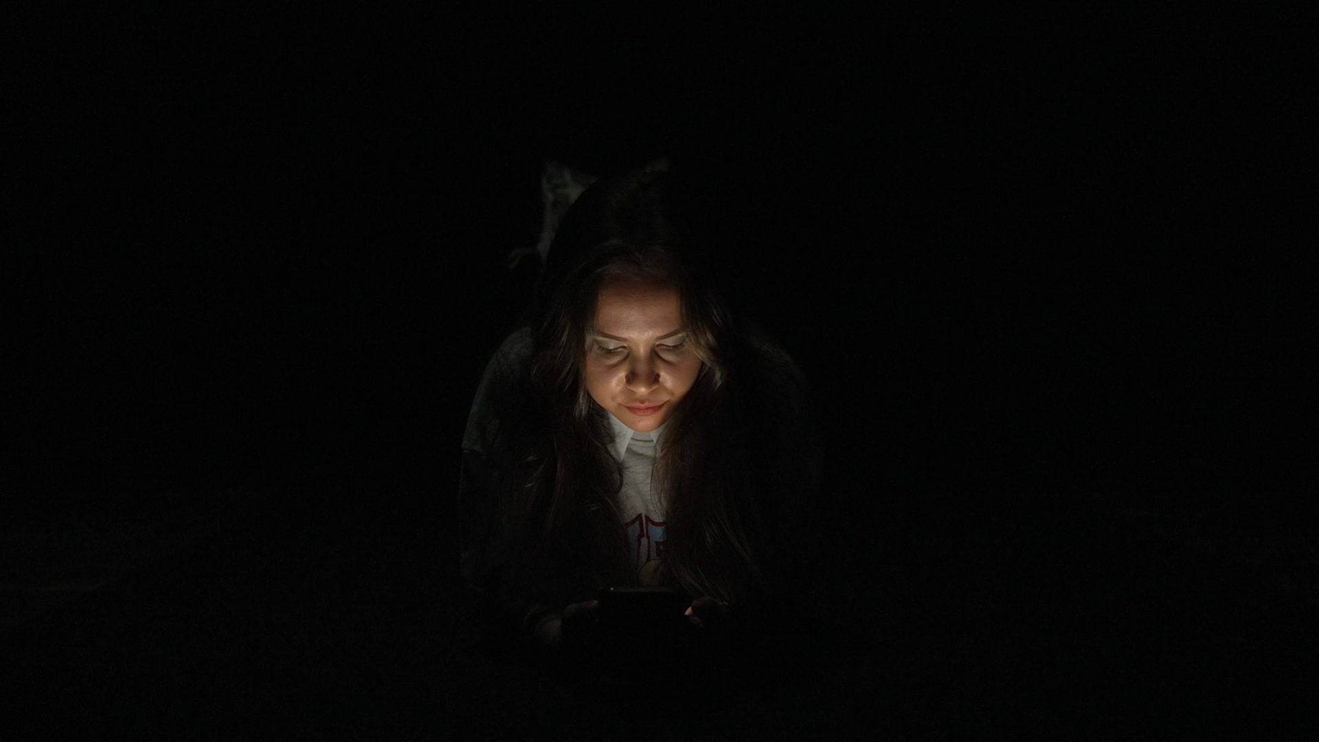 W ciemnym pomieszczeniu na podłodze leży dziewczyna. W dłoniach trzyma telefon. Ekran telefonu oświetla jej twarz.
